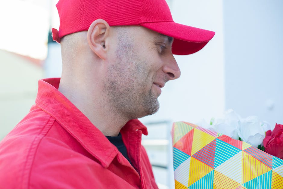 A man wearing a red cap and red jacket is holding a vibrant, patterned gift bag filled with fresh, colorful flowers, including white roses and red blooms, in a bright outdoor setting with soft natural