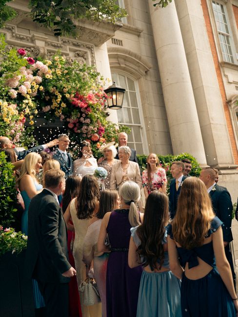 A group of elegantly dressed guests gathered outdoors on the steps of a stately building, decorated with a lavish floral arch featuring abundant pink, white, and blush roses, complemented by greenery,