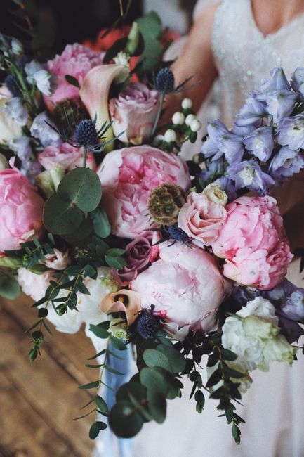 A close-up of a wedding floral arrangement featuring large pink peonies, light purple delphiniums, white roses, and sprigs of eucalyptus with rounded green leaves. The bouquet is lush and densely comp