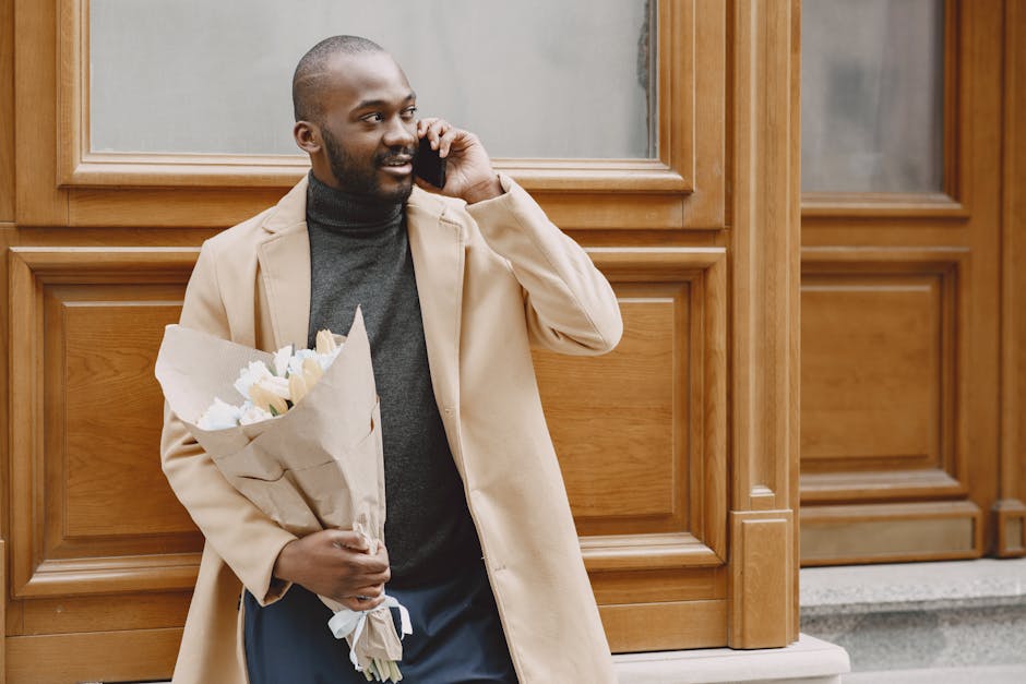 A man with dark skin and a neatly trimmed beard stands outdoors against a wooden panelled background, holding a bouquet of fresh white and cream flowers wrapped in brown paper. He is casually dressed