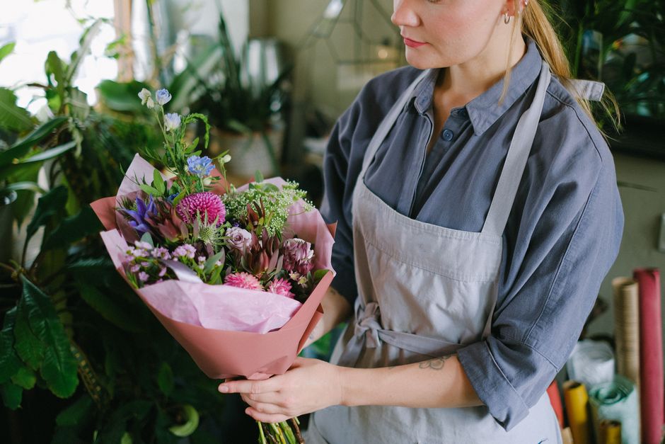 A woman wearing a gray shirt and a white apron holds a vibrant floral bouquet wrapped in pink tissue paper. The arrangement features pink carnations, purple lisianthus, blue delphiniums, and white fil